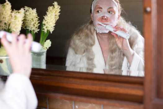 a middle-aged woman in a refreshing mask is busy with morning hygiene, brushing her teeth with an electric toothbrush. High quality photo