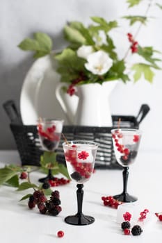 transparent gin and tonic with ice and fresh red currants and blackberries in beautiful crystal glasses against the background of a vase of flowers. High quality photo