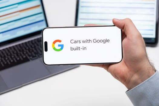 Cars with Google built in application logo on the screen of smart phone in mans hand, laptop and tablet are on the table in the background, December 2023, Prague, Czech Republic.