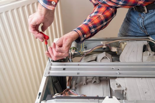 middle-aged master in a checkered suit unscrews the lid of a washing machine with a screwdriver for repair, repairs washing machines and household appliances at home, high quality photo