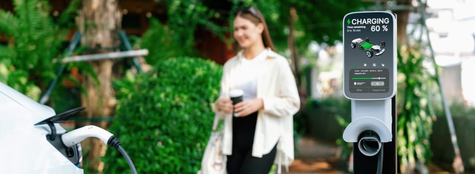 Young woman with coffee cup and sustainable urban commute with EV electric car recharging at outdoor coffee cafe in springtime garden, green city and environmental friendly EV car. Panorama Expedient