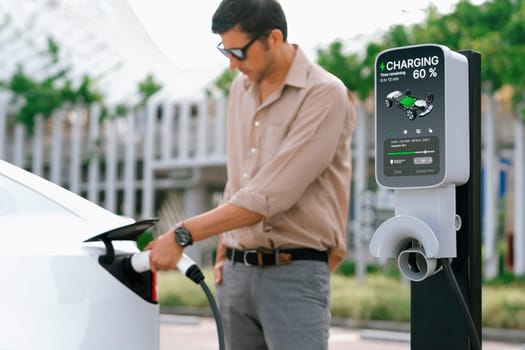 Young man put EV charger to recharge electric car's battery from charging station in city commercial parking lot. Rechargeable EV car for sustainable environmental friendly urban travel. Expedient