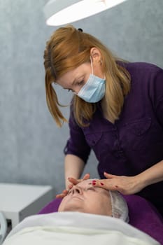 The beautician gently applies her hands to her client's cheeks. The woman is lying on a cosmetic bed and is covered with a white coverlet. On her head is a cap to secure her hair. The beautician massages her face.
