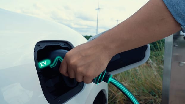 Hand insert EV charger and recharge electric car from charging station with wind turbine generator background. Smart eco-friendly EV car. Technological advancement of alternative energy. Peruse