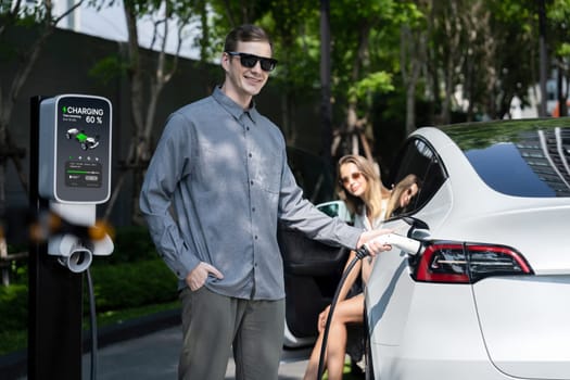 Young couple travel with EV electric car charging in green sustainable city outdoor garden in summer shows urban sustainability lifestyle by green clean rechargeable energy of electric vehicle innards