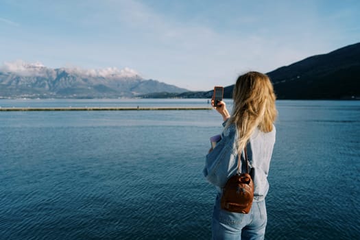 Woman shoots a sea and a mountain range with a smartphone while standing on the shore. Back view. High quality photo