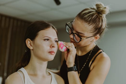 One young handsome Caucasian makeup artist applies foundation with a pink sponge to the face of a girl sitting in a chair early in the morning in a beauty salon, close-up side view. Step by step.