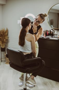 One young handsome Caucasian makeup artist applies natural shadows with a brush to the eyelids of a girl sitting in a stylish chair early in the morning in a beauty salon, close-up side view. Step by step.