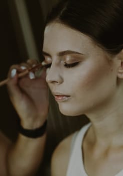 Portrait of a young beautiful Caucasian girl sitting on a chair with closed eyes in a beauty salon, where the master is applying concealer with a brush on her face, side view.