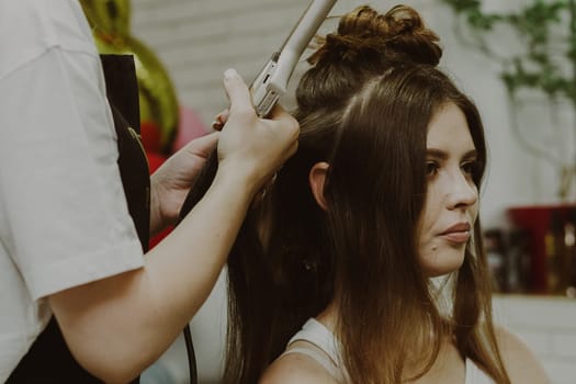 Portrait of a young Caucasian girl sitting sideways in a hairdresser's shop, where the master is curling her hair on a flat iron, close-up view.