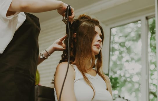 Portrait of a young beautiful brunette Caucasian girl sitting in a hairdresser's shop, where the master is curling her long hair on a flat iron, close-up view from below.