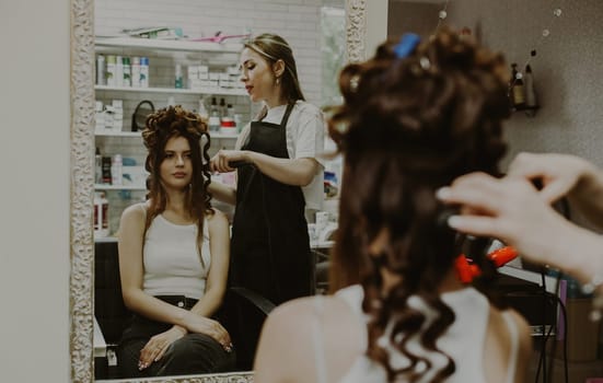 Portrait of a beautiful young brunette girl sitting in a chair, reflected in the mirror, which the hairdresser twists her hair on a curling iron, doing a wedding hairstyle, close-up top view.