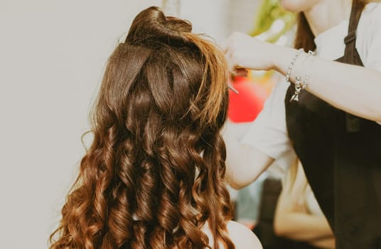 Portrait of a young beautiful Caucasian brunette girl sitting with pina in a hairdresser's shop, where the master is doing her hairstyle of tousled curls, close-up view from the side.