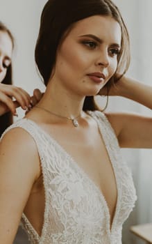 Portrait of a beautiful brunette bride who's bridesmaid is fastening the clasp of a necklace around her neck, standing in a room by a window, close-up side view.
