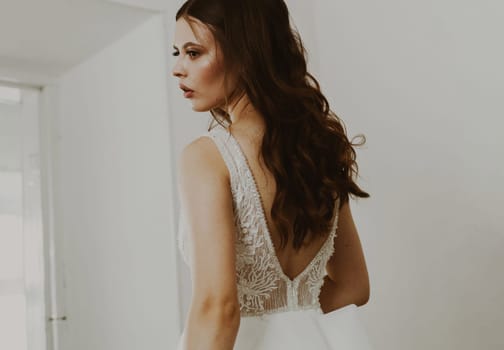 Portrait of a beautiful brunette bride in a white wedding dress standing in a room looking at herself in a mirror.
