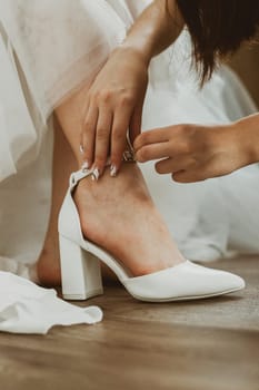 The hand of an unrecognizable bridesmaid helps to put white wedding shoes on the bride's leg, close-up side view.