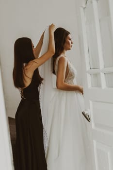 A beautiful young bridesmaid helps the bride put on a white veil while standing in the room behind the door, side view, close-up.