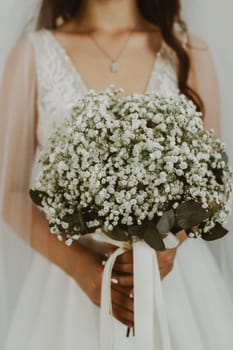 Portrait of one unrecognizable young Caucasian bride in a white dress and veil holding a wedding bouquet of white boutonnieres in her hands in front of her, side view, close-up.