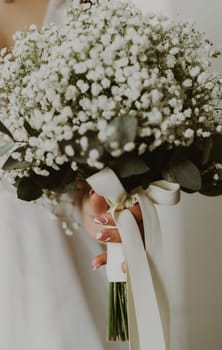 Portrait of one unrecognizable young Caucasian bride holding in her hands with a beautiful delicate manicure a wedding bouquet of white boutonnieres with ribbons, side view, close-up with selective focus.