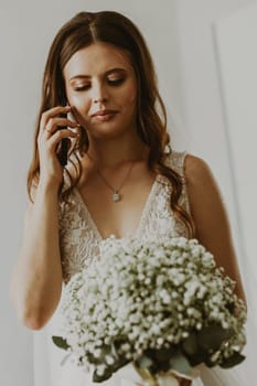 Portrait of one beautiful young Caucasian and happy brunette bride calling her groom on a mobile phone in the room during the day, close-up side view.