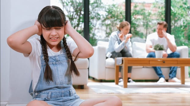 Stressed and unhappy young girl huddle in corner, cover her ears blocking sound of her parent arguing in background. Domestic violence at home and traumatic childhood develop to depression. Synchronos