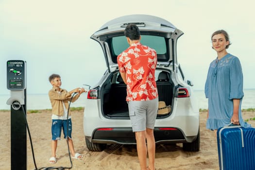 Family vacation trip traveling by the beach with electric car, lovely family taking luggage out while charging EV car battery with clean energy. Alternative family travel by eco-friendly car.Perpetual