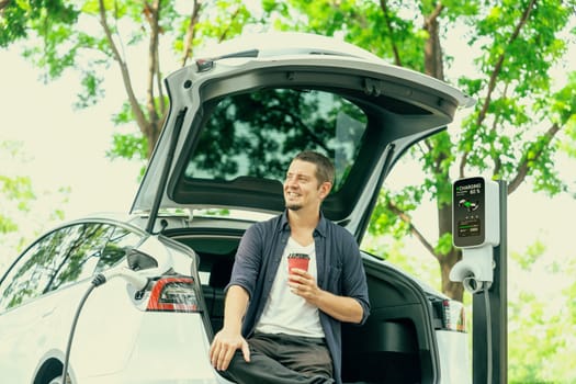 Man recharging battery for electric car during road trip travel EV car in natural forest or national park. Eco friendly travel during vacation and holiday. Exalt