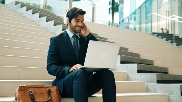 Professional business man sitting at stairs while working on laptop. Skilled project manager listening music from headphone and checking email and discussion about marketing plan. Outdoor. Exultant.