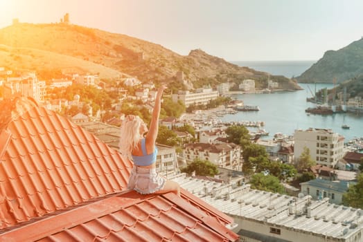 Woman sits on rooftop with outstretched arms, enjoys town view and sea mountains. Peaceful rooftop relaxation. Below her, there is a town with several boats visible in the water.