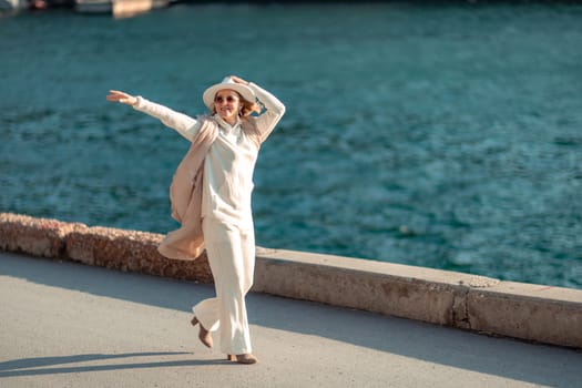 Happy blonde woman in a white suit and hat posing at the camera against the backdrop of the sea.