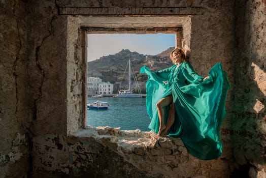 Rear view of a happy blonde woman in a long mint dress posing against the backdrop of the sea in an old building with columns. Girl in nature against the blue sky