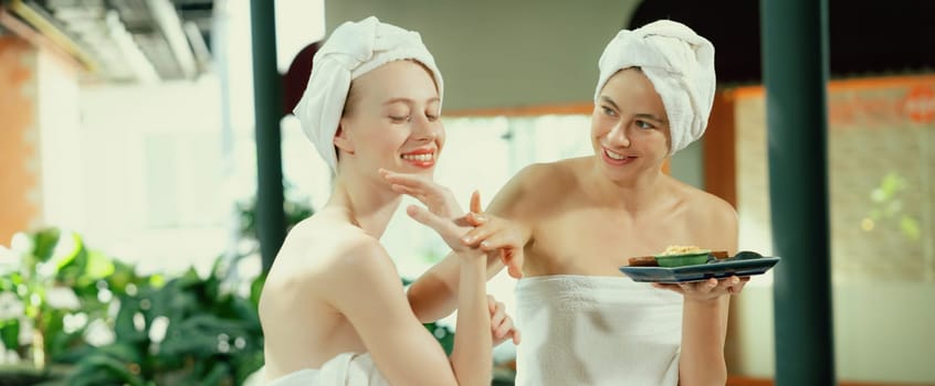 Portrait of two attractive woman in towel giggling during hold the herbal bowl surrounded by natural spa environment. Pretty girls with beautiful skin using herbal scrub at spa salon.Tranquility.