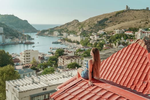 Woman sits on rooftop, enjoys town view and sea mountains. Peaceful rooftop relaxation. Below her, there is a town with several boats visible in the water. Rooftop vantage point