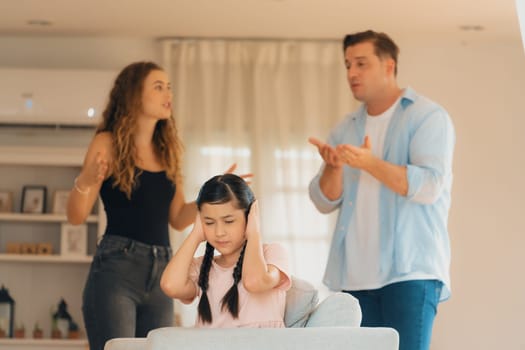 Annoyed and unhappy young girl sitting on sofa trapped in middle of tension by her parent argument in living room. Unhealthy domestic lifestyle and traumatic childhood develop to depression.Synchronos