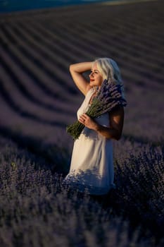 Blonde woman poses in lavender field at sunset. Happy woman in white dress holds lavender bouquet. Aromatherapy concept, lavender oil, photo session in lavender.