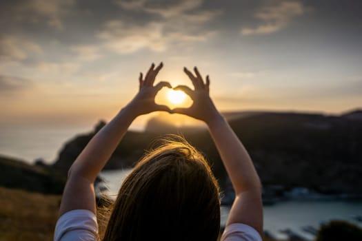 Happy woman on mountain peak, she makes heart shape with hands. Mountain, overlooking sea at sunset. Depicting love shape amidst scenic natural setting
