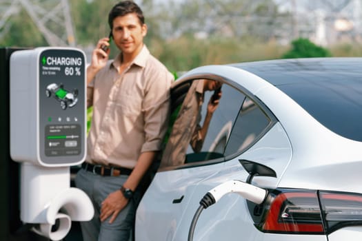 Man pay for electricity with smartphone while recharge EV car battery at charging station connected to power grid tower electrical as electrical industry for eco friendly car utilization.Expedient