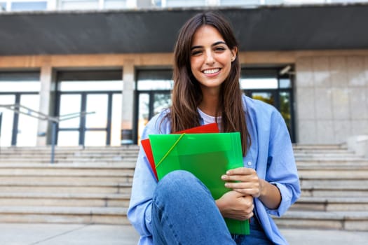 Happy, smiling female college student looking at camera holding folders sitting outside university building.Education concept.