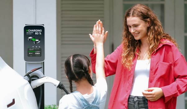 Happy little young girl learn about eco-friendly and energy sustainability as she help her mother recharge electric vehicle from home EV charging station. EV car and modern family. Panorama Synchronos