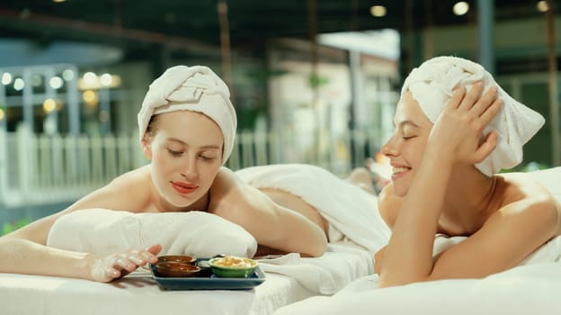 Couple of beautiful young girls lie on spa bed during interested in homemade beauty facial mask. Attractive woman touching herbal facial mask. Surrounded with nature environment. Tranquility.