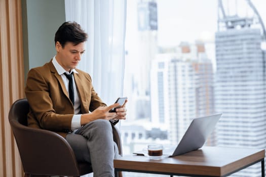 Closeup of handsome businessman using mobile phone while sitting near window with skyscraper view. Professional male leader taking a photo marketing, business project displayed on laptop. Ornamented.