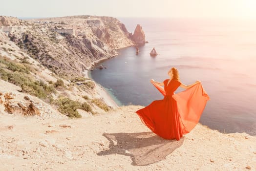 Side view a Young beautiful sensual woman in a red long dress posing on a rock high above the sea during sunrise. Girl on the nature on blue sky background. Fashion photo.