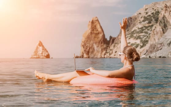 Woman freelancer works on laptop swimming in sea on pink inflatable ring. Pretty lady typing on computer while floating in the sea on inflatable donut at sunset. Freelance, remote work on vacation