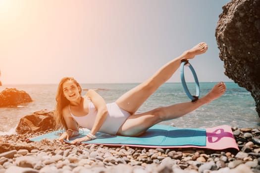 Young woman with black hair, fitness instructor in pink sports leggings and tops, doing pilates on yoga mat with magic pilates ring by the sea on the beach. Female fitness daily yoga concept