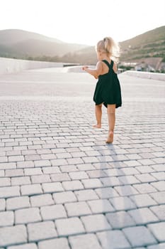 Little girl walks barefoot along a tiled road at the foot of the mountains, waving her arms. Back view. High quality photo