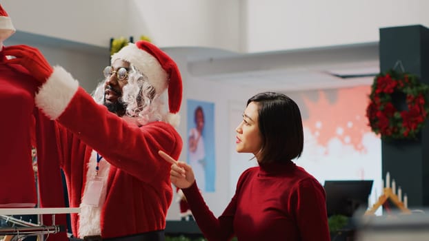 Retail assistant impersonating Santa Claus helping customer in shopping store find red blouse needed for Christmas party outfit. Worker assisting woman in fashion shop during festive season