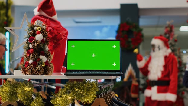 Mockup laptop on xmas decorated clothing store display table showing information about products. Green screen device in shopping mall fashion shop useful for looking up clothes prices