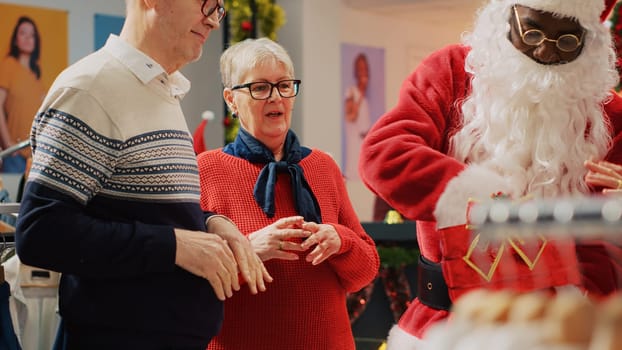 Clients participating in Christmas raffle hold by retail assistant dressed as Santa Claus in festive ornate clothing store. Lucky elderly couple excited after winning promotional fashion shop contest