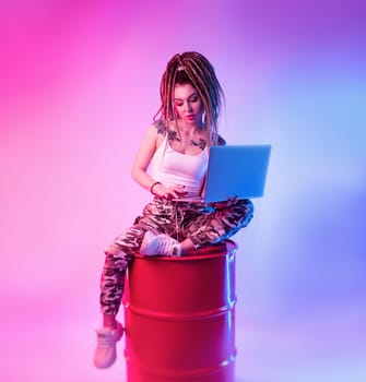 sexy girl with braids dreadlocks on her head in neon light with a laptop on a light background copy paste