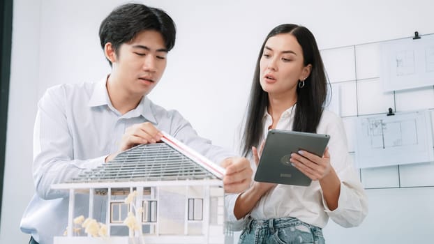 Beautiful young caucasian project manager tells asian engineer about building detail by using tablet while engineer measuring house model on meeting table with blueprint scatter around. Immaculate